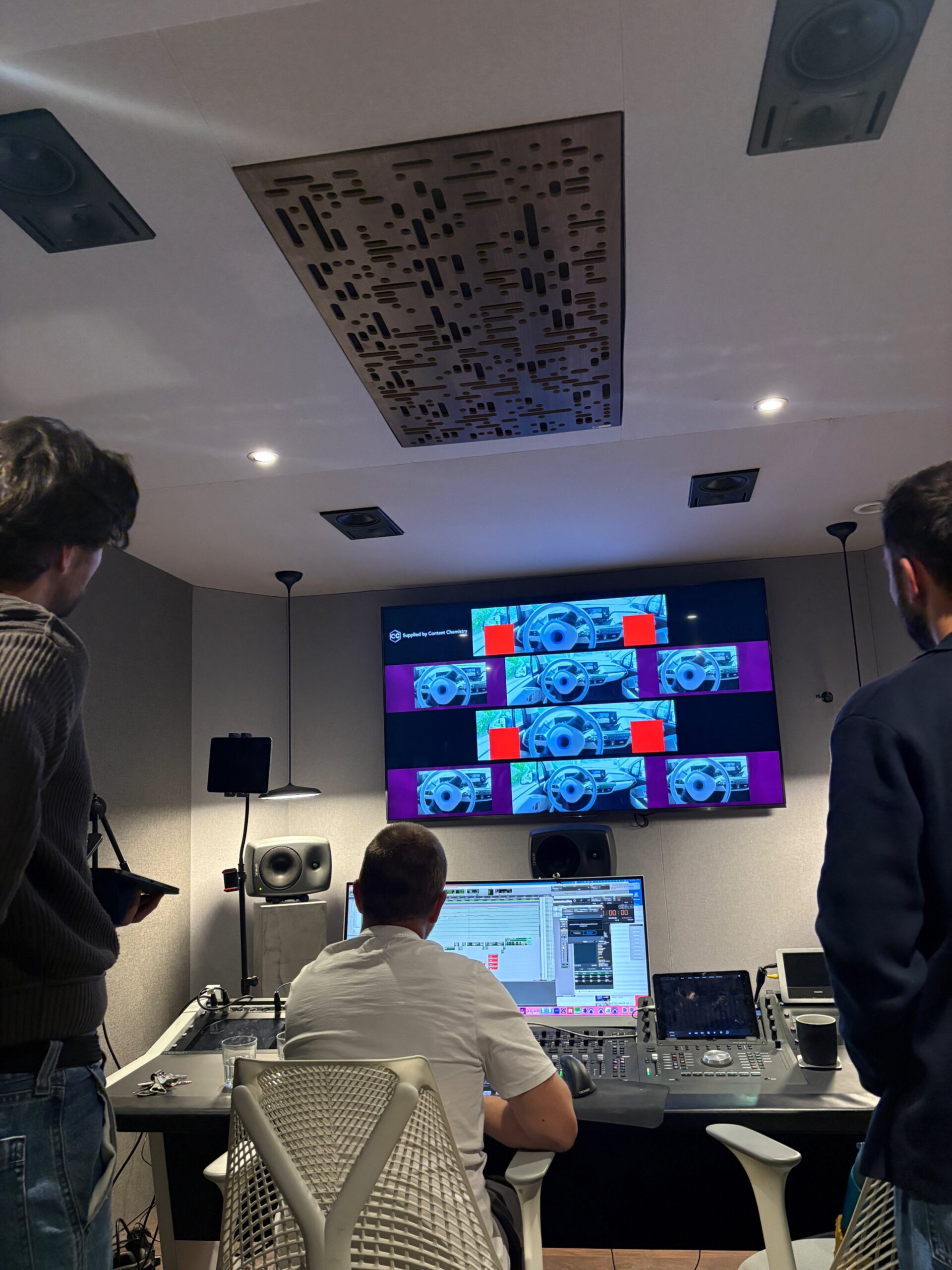 Three people in a sound editing suite reviewing visuals on multiple screens and audio software on a mixing console.