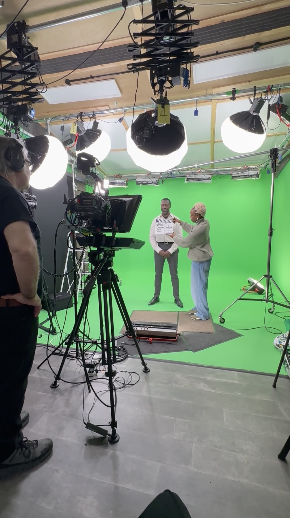 Two presenters standing in front of a green screen in a video production studio, surrounded by lighting equipment and cameras.